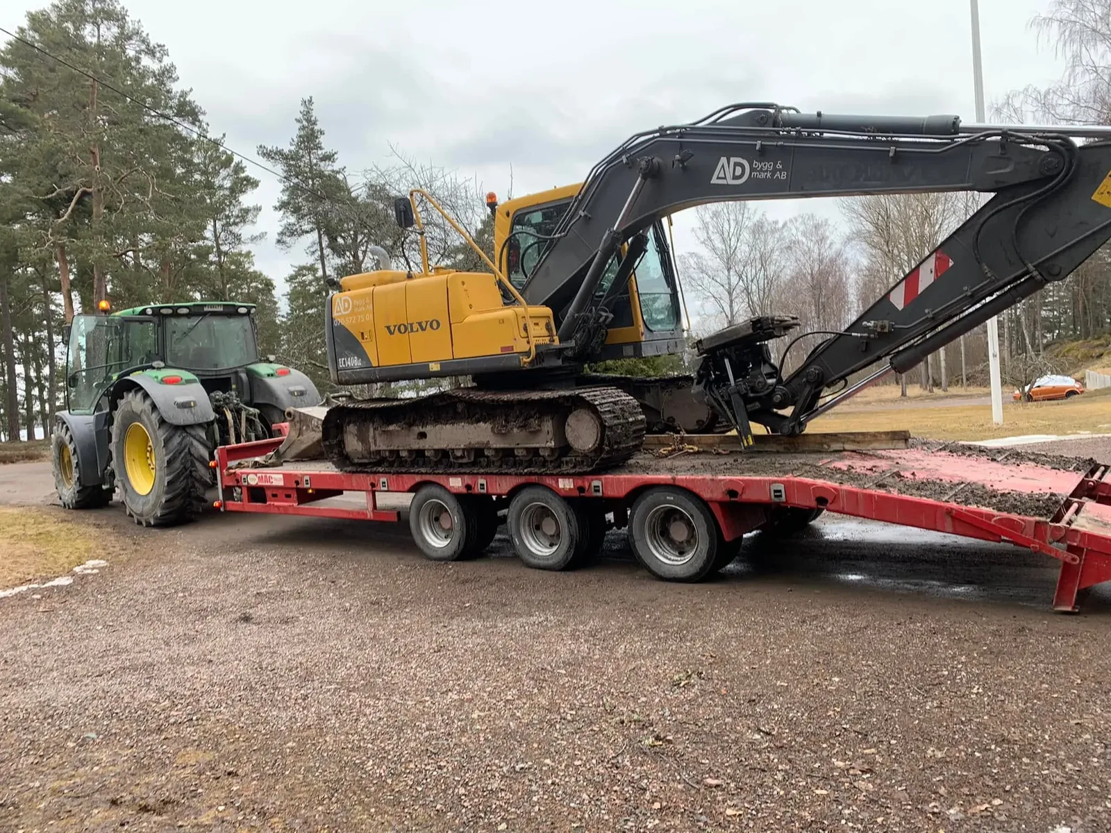 Grävmaskin med AD Bygg & Mark-logga på bommen lastad på en röd maskintrailer bakom en grön traktor