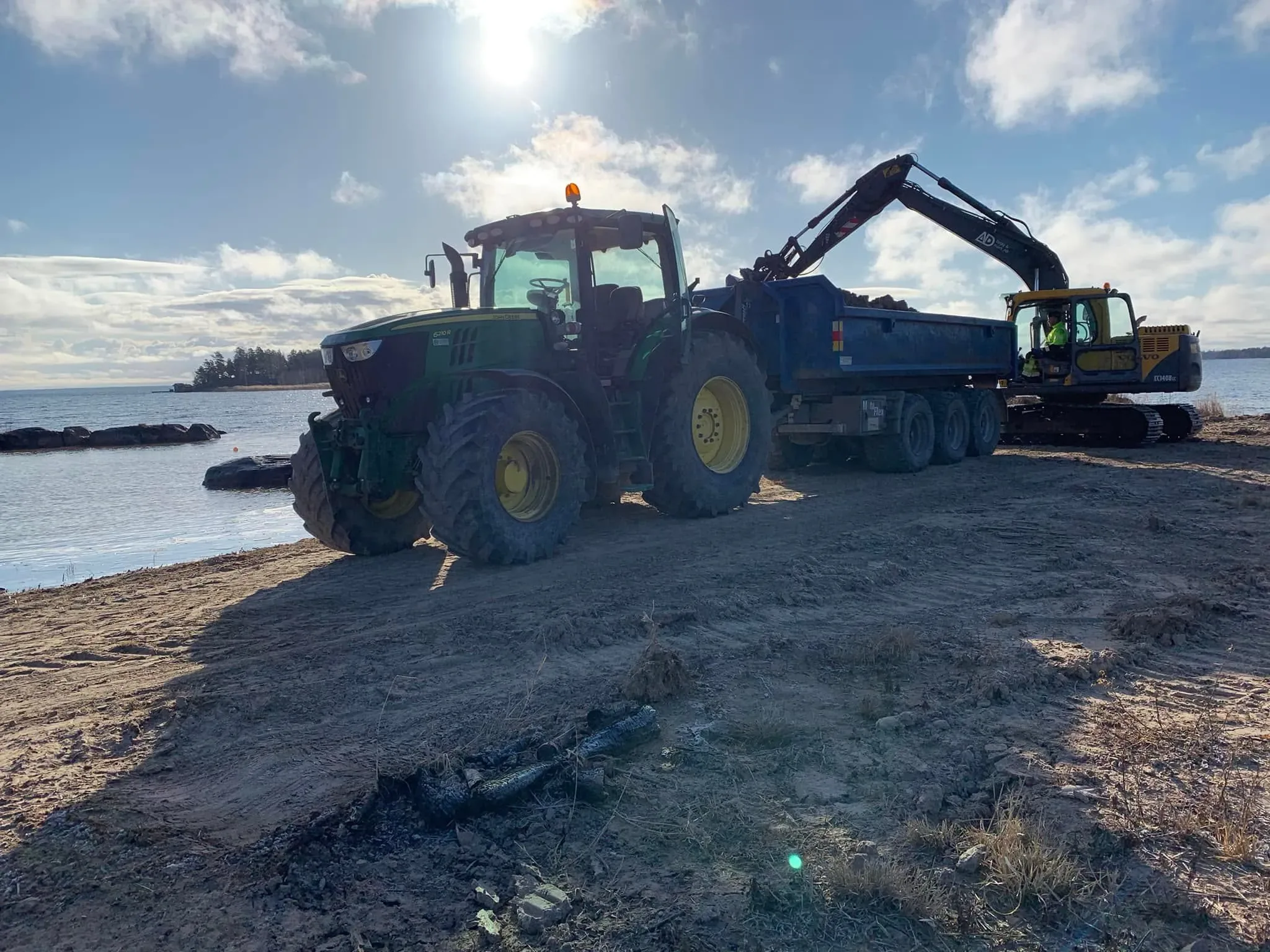 Grävmaskin med AD Bygg & Mark-logga lastar jordmassor i en dumpervagn bakom en grön traktor vid en sjöstrand i Värmland