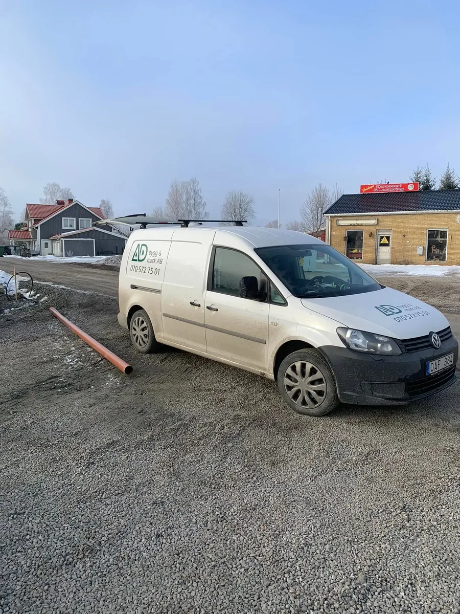 Vit servicebil med AD Bygg & Mark-logga och telefonnummer på dörren parkerad på en grusplan