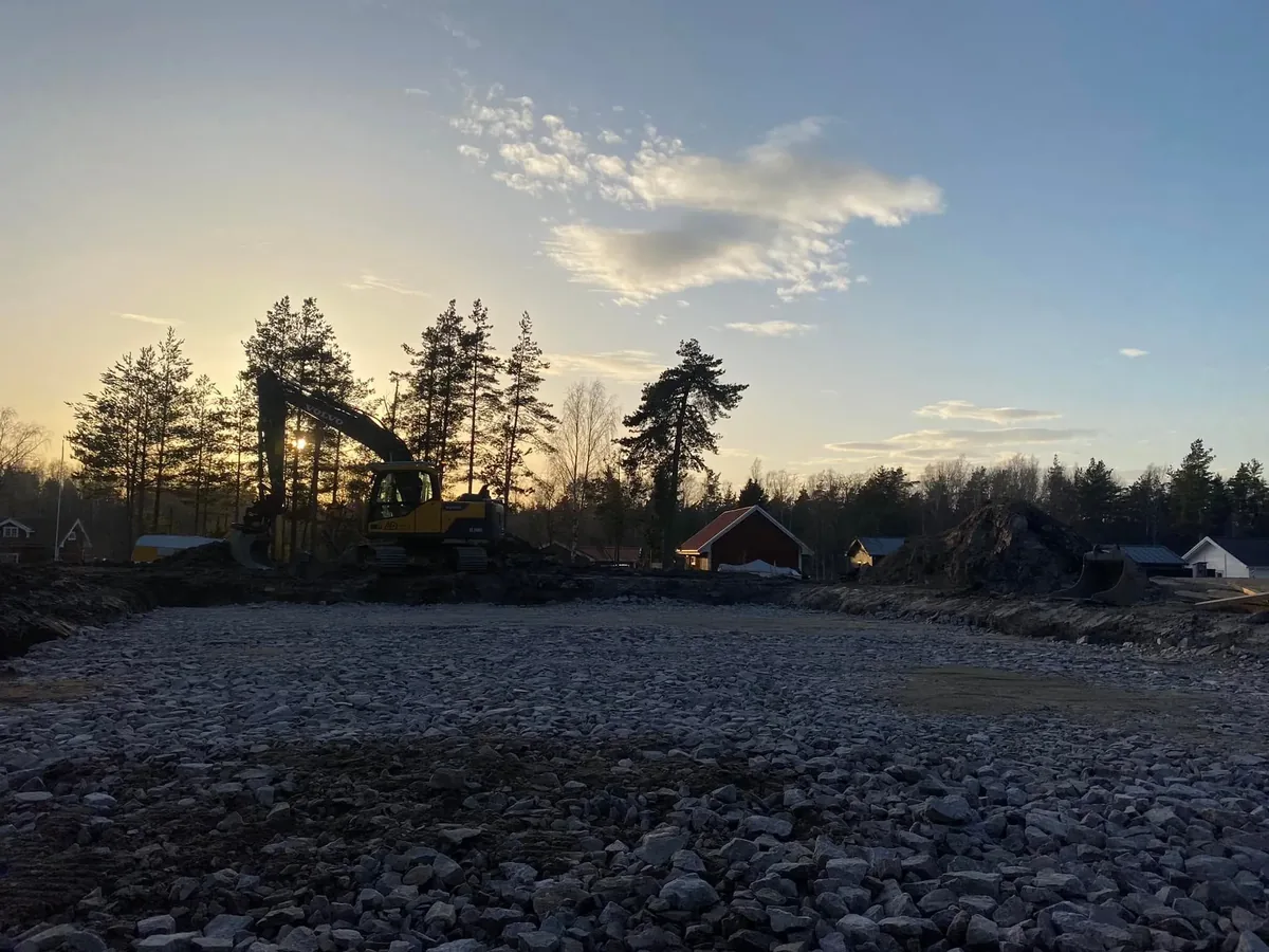 Stenig tomt som förbereds för bygge, med en grävmaskin i motljus