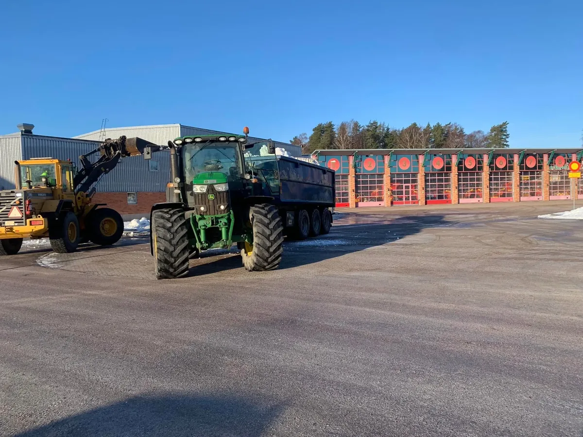 Grön traktor med dumpervagn och en gul lastare på en asfalterad gård
